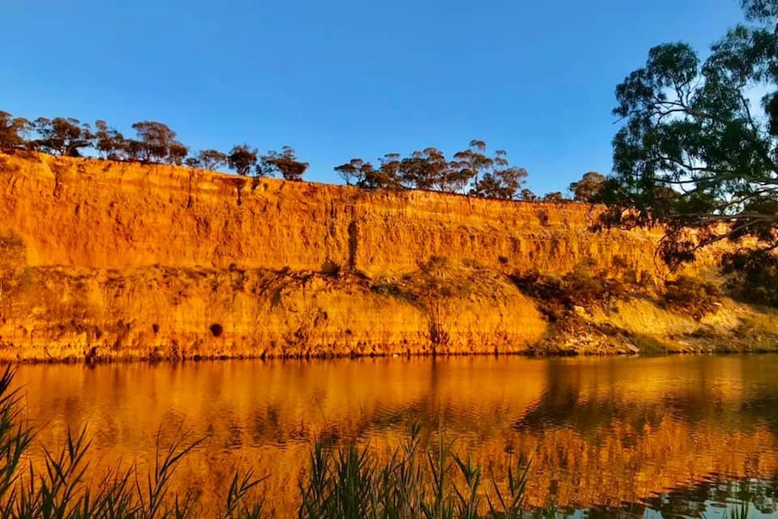Red cliffs shown in full sunlight, with a reflection in the river below, under a blue sky.