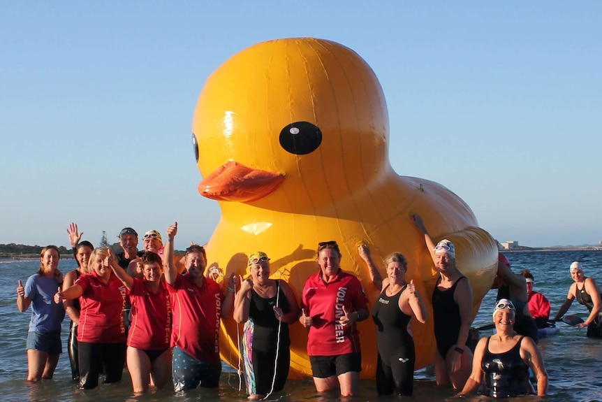 A community swimming group stands in the ocean with a giant rubber duck mascot.