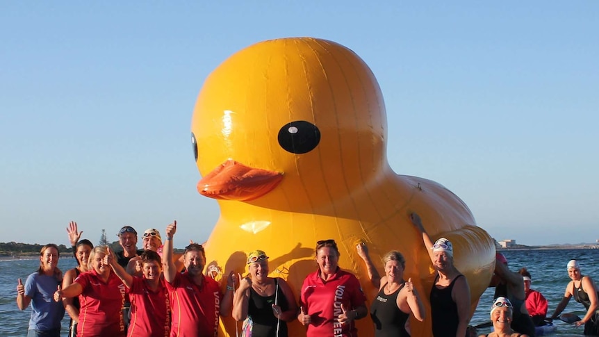 A community swimming group stands in the ocean with a giant rubber duck mascot.