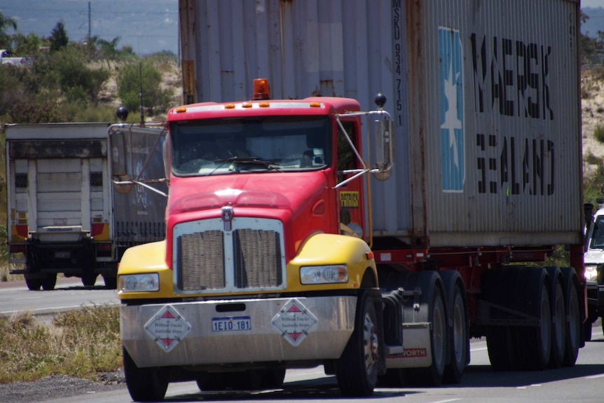 A red and yellow truck can towing a trailer with a sea container on it travels along Roe Highway.