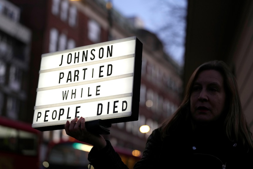 A woman holding a sign in a dimly lit street, which reads "Johnson partied while people died"