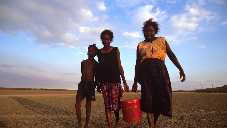 Two women and a boy carrying a bucket of water in the desert.