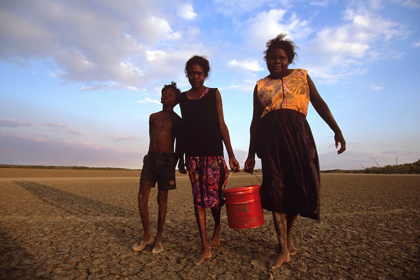 Two women and a boy carrying a bucket of water in the desert.