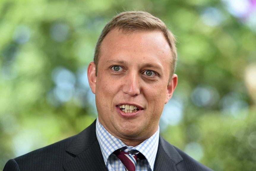 A fair-haired man approaching middle age, sharply dressed in a suit and standing in front of some trees.