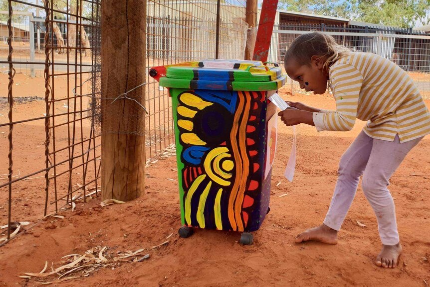 A child reads a sign tacked to the front of a bin painted with bright colours.