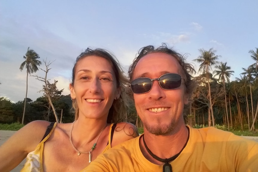A woman and man pose for a selfie in front of palm trees. 