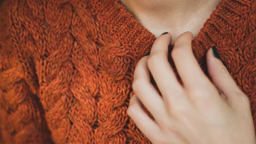 A woman wearing a red knit jumper holds her hand to her collarbone.
