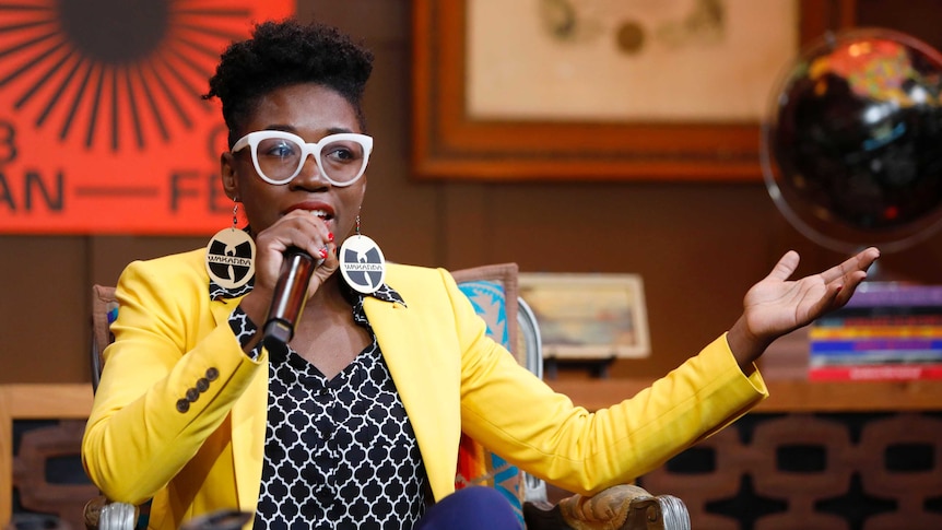 African-American woman wearing white glasses, yellow blaze and Wakanda earrings speaks into a microphone she holds in one hand.