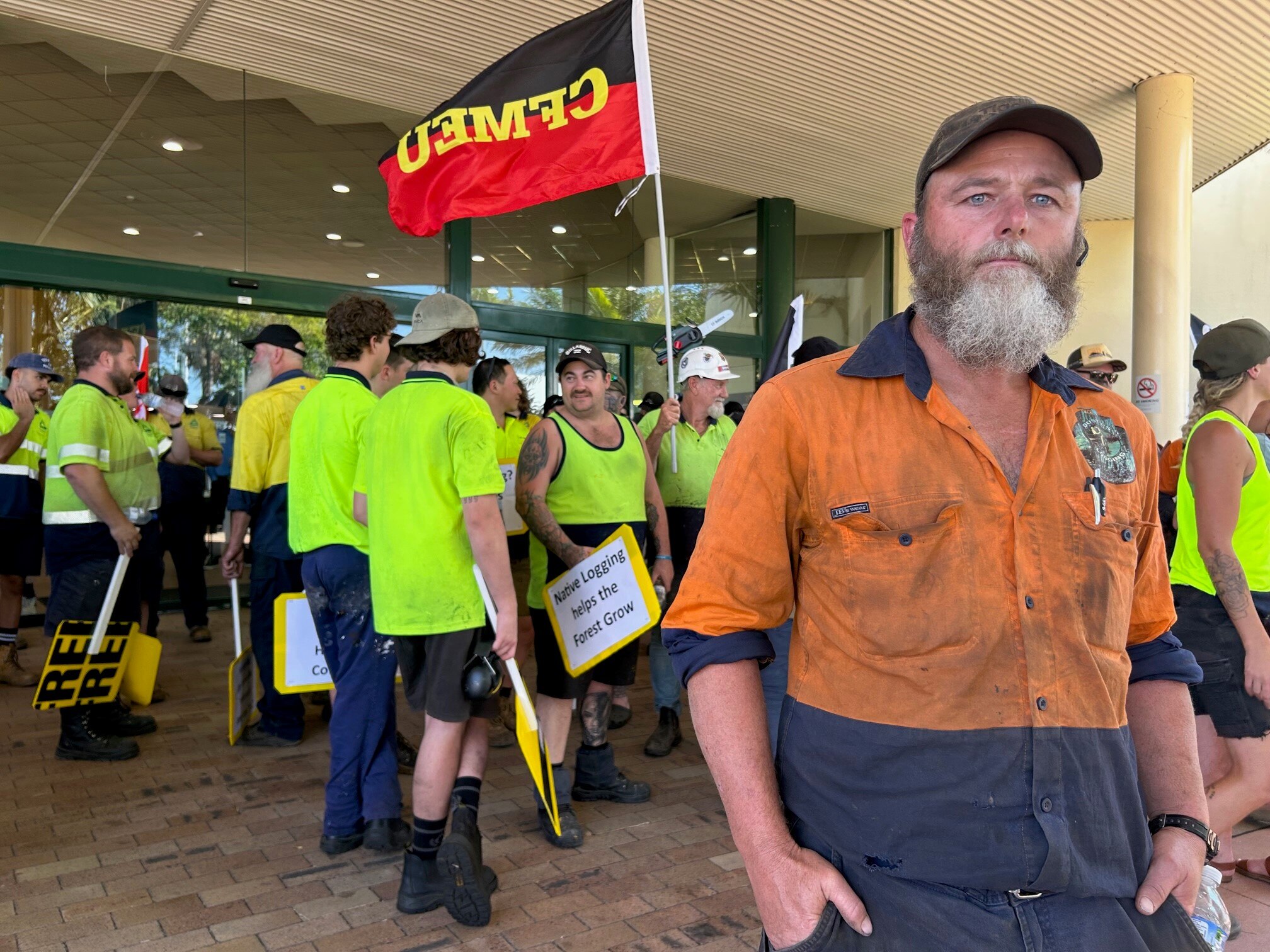 Bearded man in high-vis gear stand in front of protesters.