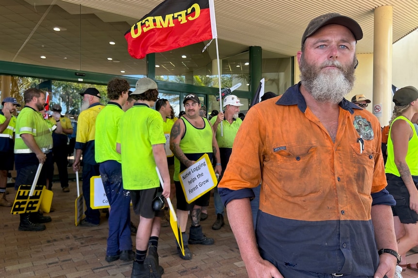 Bearded man in high-vis gear stand in front of protesters.