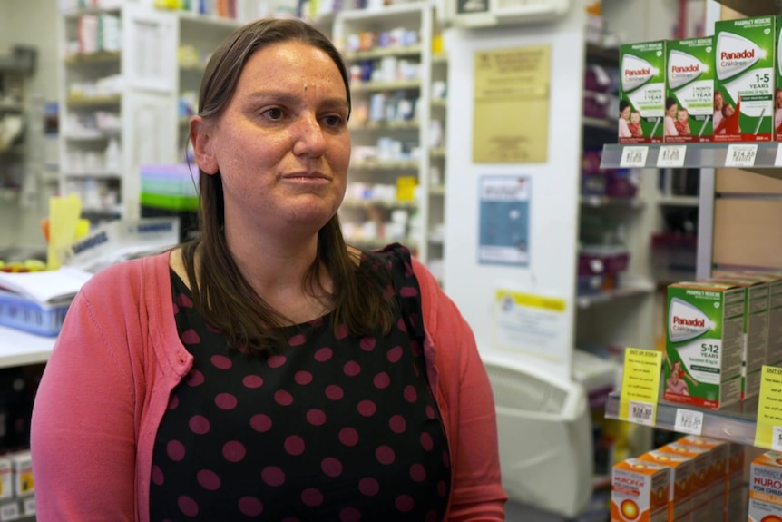 Femme debout dans sa pharmacie portant un haut imprimé et un cardigan rose.