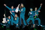 Six-piece band Daði & Gagnamagnið are seen in matching blue tracksuits against a black backdrop. Some hold synth instruments.