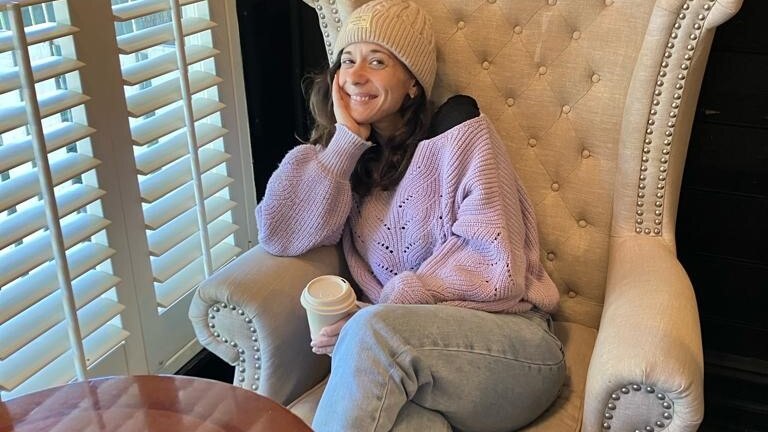 young woman sitting in a chair smiling at the camera wearing a beanie and jumper