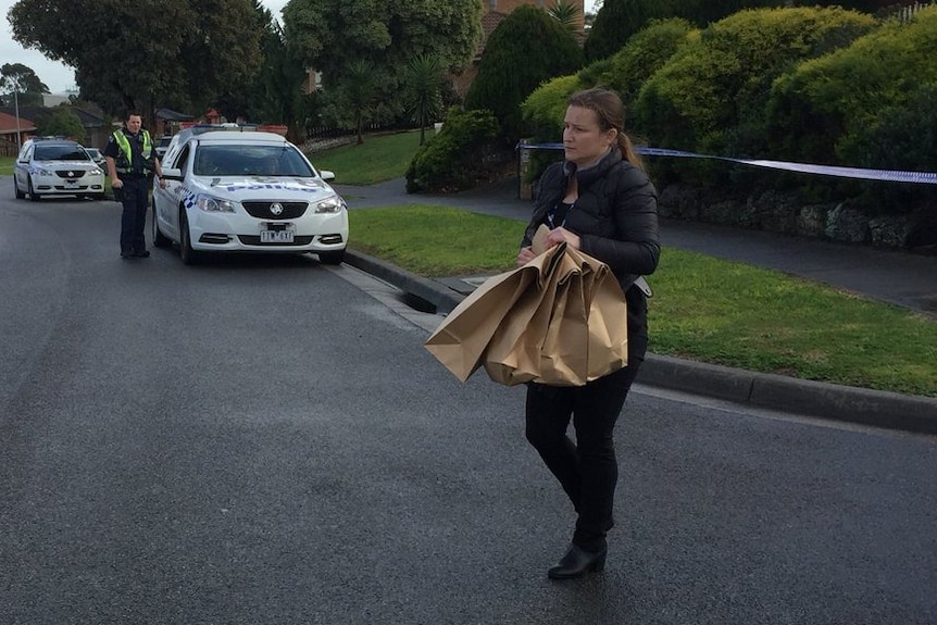 A police woman carries brown paper bags from the house in Cranbourne North while a police officer looks on in the background.