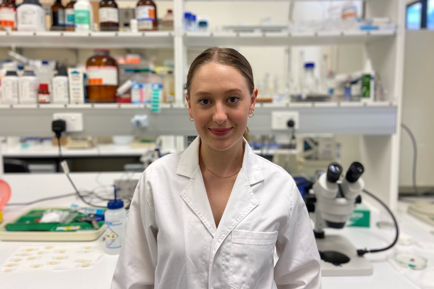 A white, blonde woman looks to the camera and slightly smiles as she stands in a lab, wearing a lab coat.