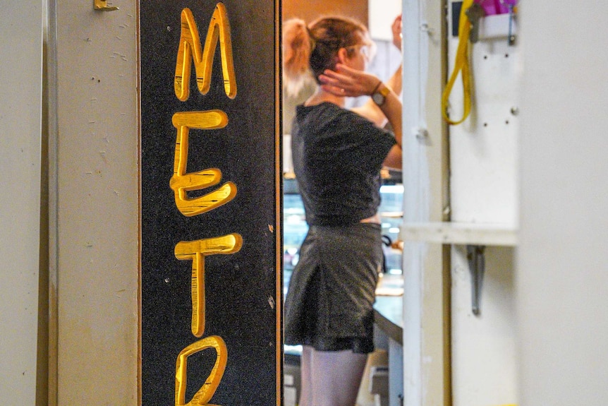 A woman wearing a black t-shirt, skirt and apron is seen through a narrow doorway.