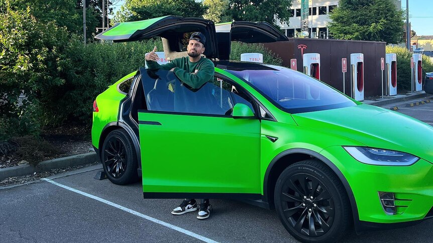 A man wearing a car backwards stands by a bright green car, with its doors open.