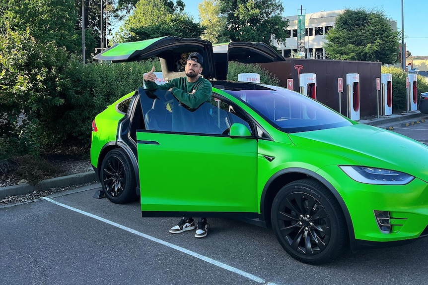 A man wearing a car backwards stands by a bright green car, with its doors open.