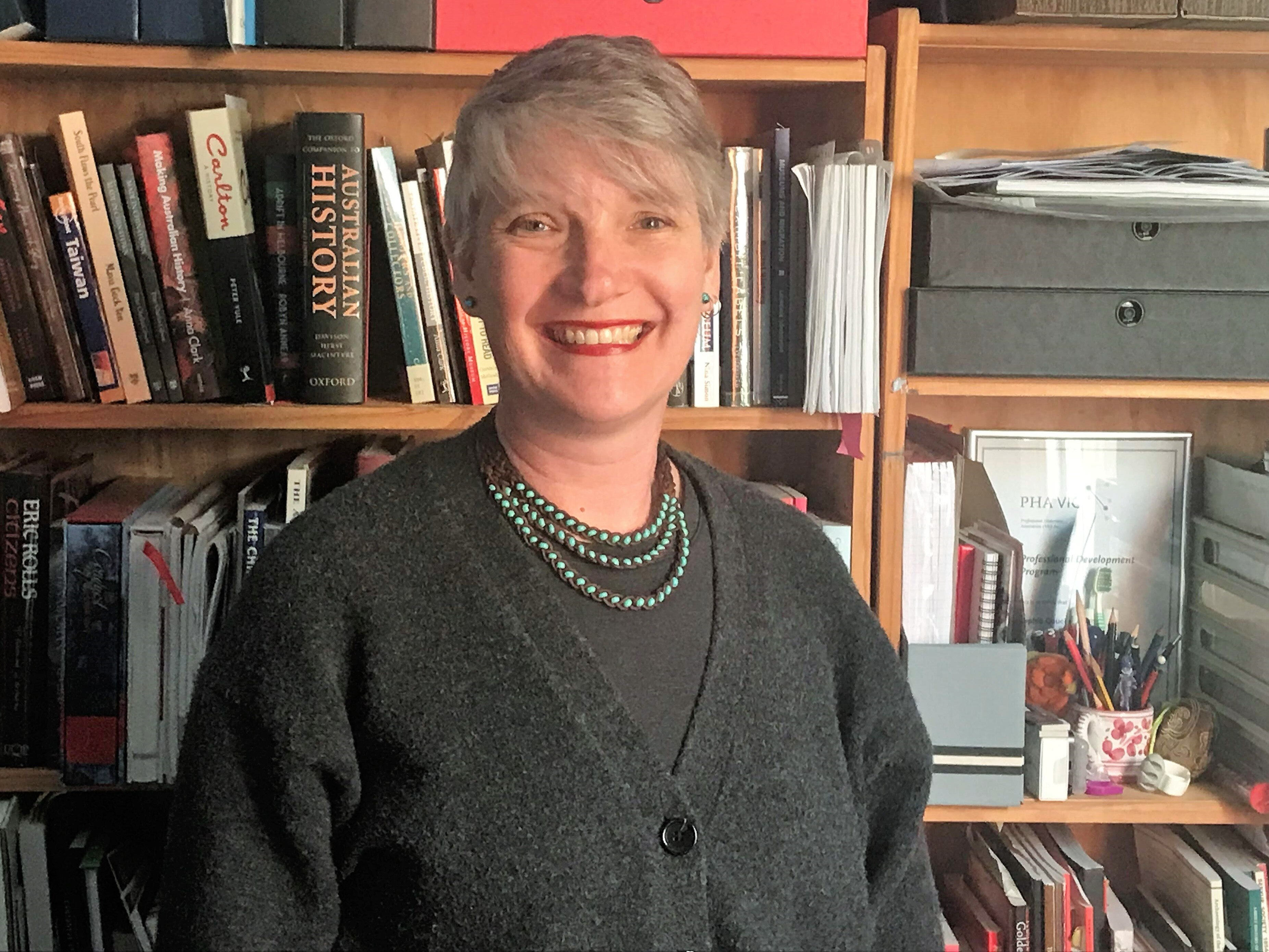 A woman with blonde hair wearing a charcoal cardigan and a green bead necklace in front of a bookshelf.