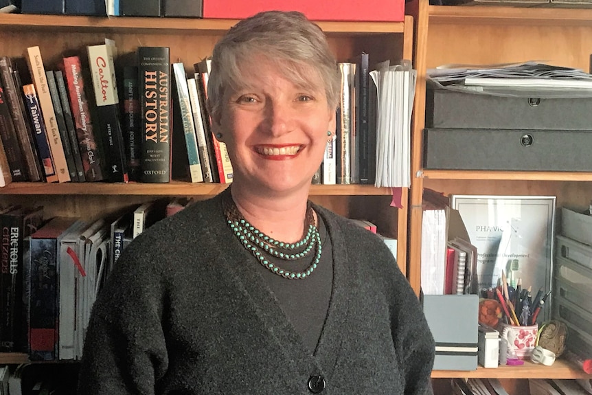 A woman with blonde hair wearing a charcoal cardigan and a green bead necklace in front of a bookshelf.