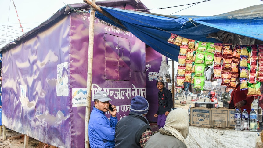 A woman stands outside a purple tent while a man talks on a mobile phone.