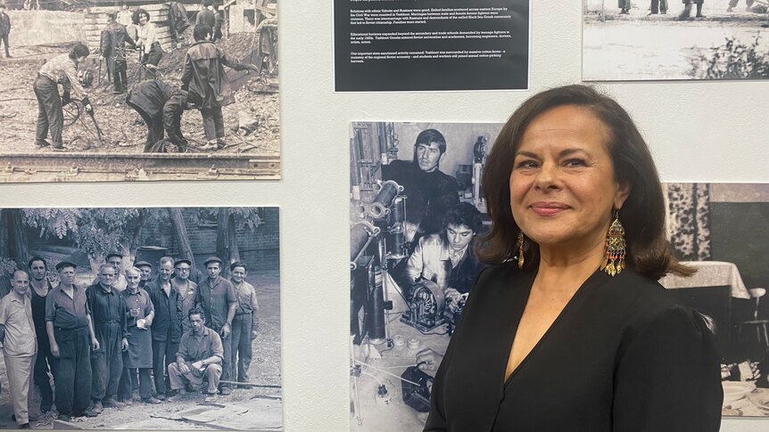 A dark-haired Greek woman in front of a wall of black and white photographs of Greek workers in the 1950s