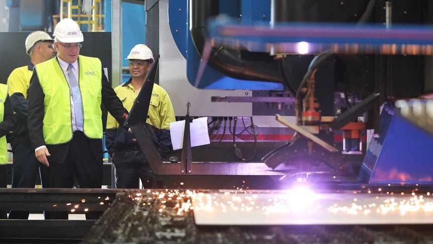 Dressed in high-vis and a hard hat, Mr Morrison looks at a sparking machine that's cutting metal