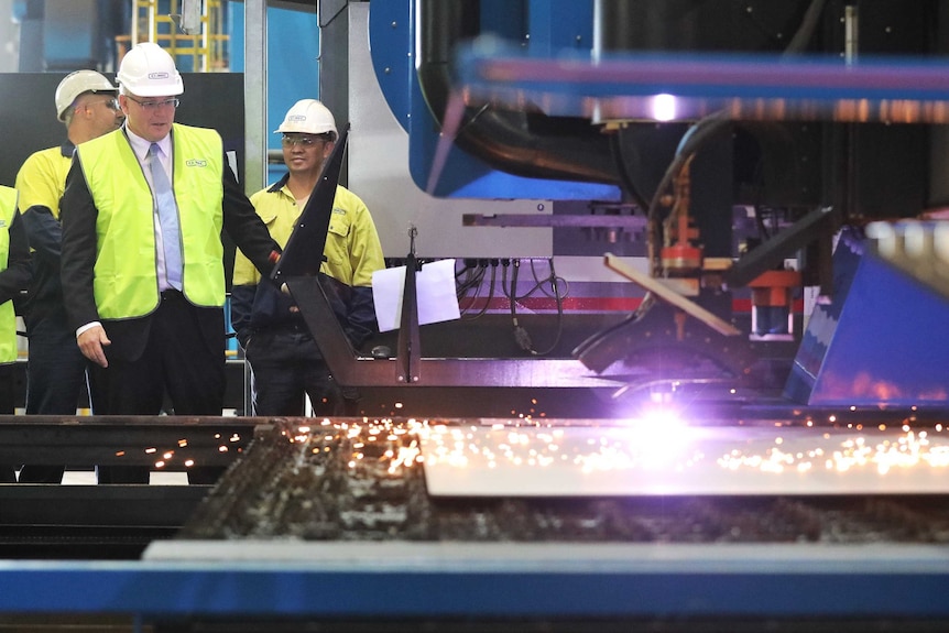 Dressed in high-vis and a hard hat, Mr Morrison looks at a sparking machine that's cutting metal