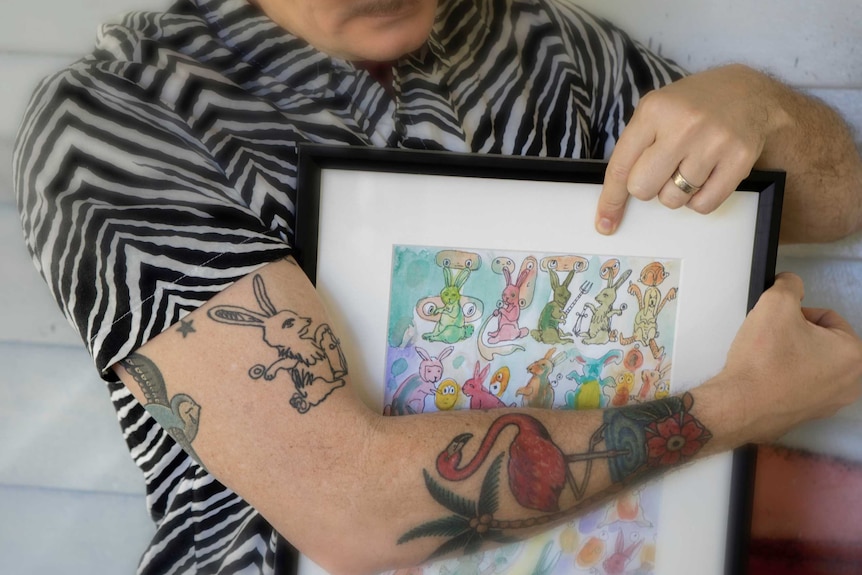 Brad holds a watercolour picture of several bunny rabbits and shows his bicep, which has a tattoo of one of the bunny rabbits.