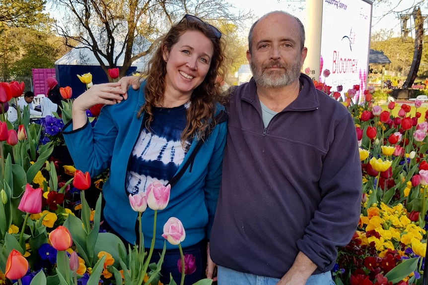 A woman and a man stand in front of colorful flowers.
