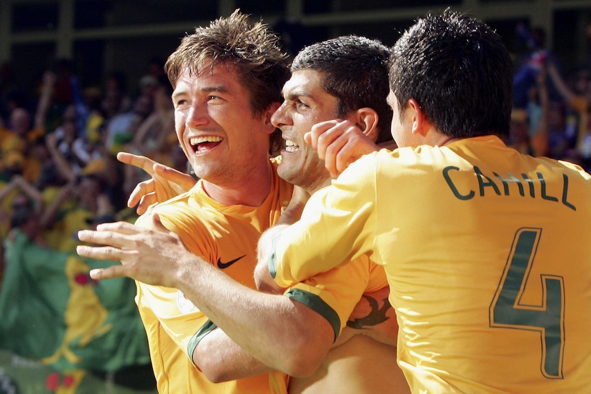 Three Socceroos embracing, two looking to their right and the third with his back to the camera. The third is Tim Cahill.