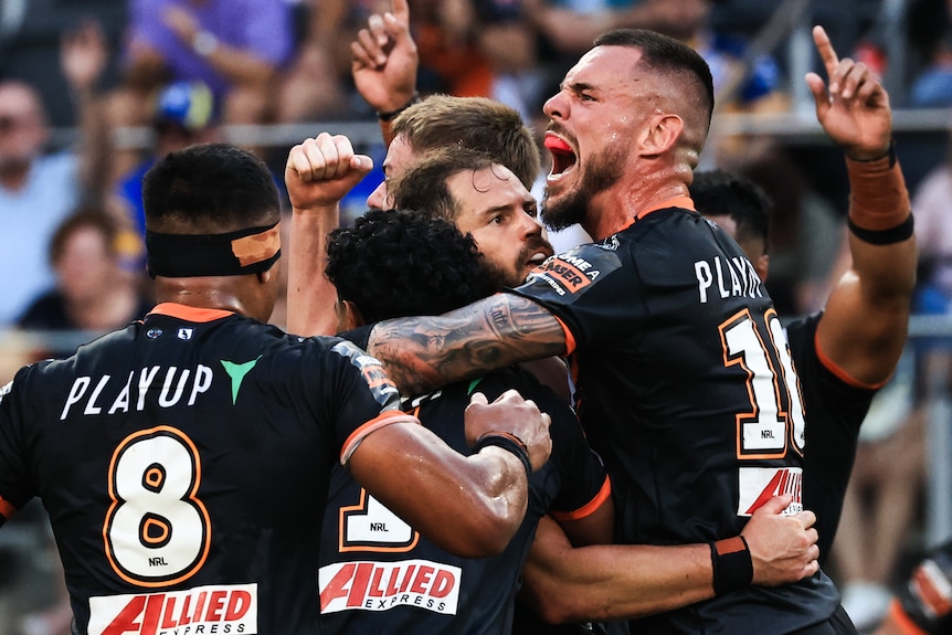 A group of Wests Tigers NRL players celebrate together as one player (number 10) roars and pumps his fist.