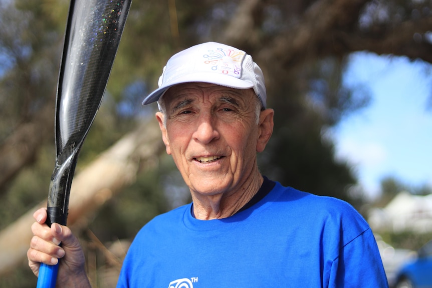 An older man in a white hat and blue shirt smile at the camera, paddle in his hand.