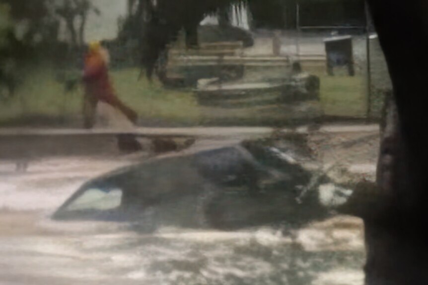 A blurry phone photo of a car swept along by brown flood water, as an SES worker in orange runs along the flood back nearby.