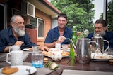 Three men sit around a table laughing while they share morning tea.