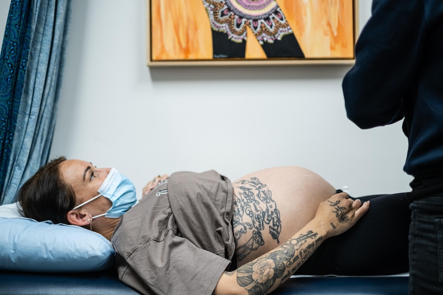 A pregnant woman wearing a facemask lays on a midwife’s bed in a women’s clinic.