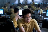 A man in a yellow shirt and dirty face on a computer with other men on computers around him. 
