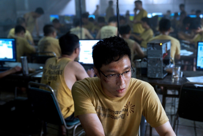 A man in a yellow shirt and dirty face on a computer with other men on computers around him. 