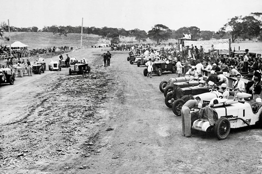 A black and white photograph showing race cars and a crowd of people watching