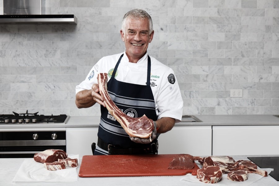 Man holds a leg of steak in a kitchen. He wears a white chef's shirt and apron and is smiling.
