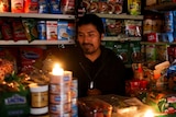 A man in a store surrounded by cans and packets of food has set up tea-light candles in the dark.