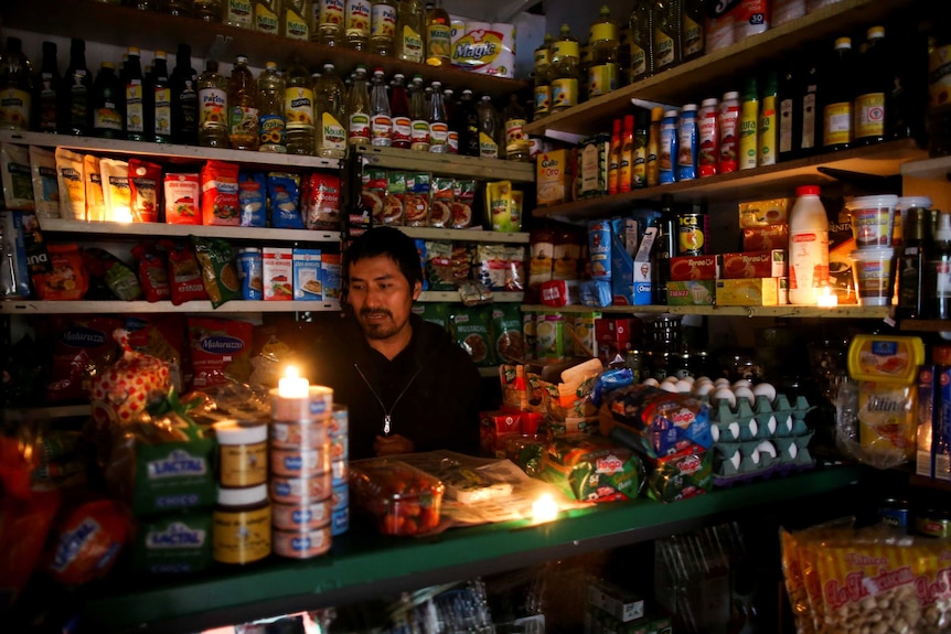 A man in a store surrounded by cans and packets of food has set up tea-light candles in the dark.