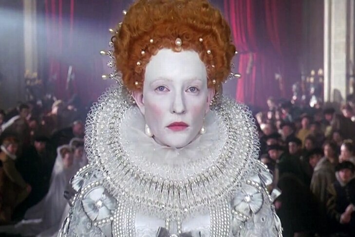 Queen Elizabeth I stands before her courtiers, in full white makeup and gown and her red wig.