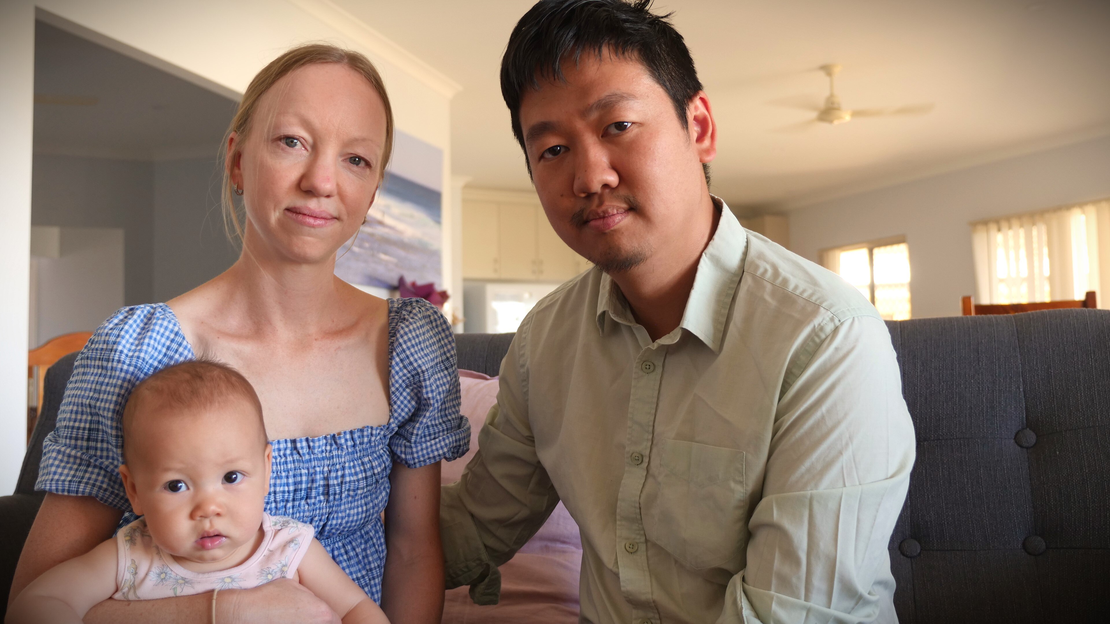 Young couple sit on grey couch holding their baby girl. 
