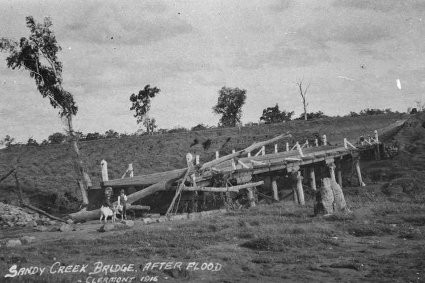 Black and white photo showing a damaged wooden bridge with two children standing in the rubble on the road.