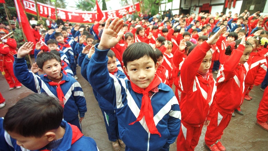 A group of young children wearing blue and red tracksuits with red ties salute towards the camera.