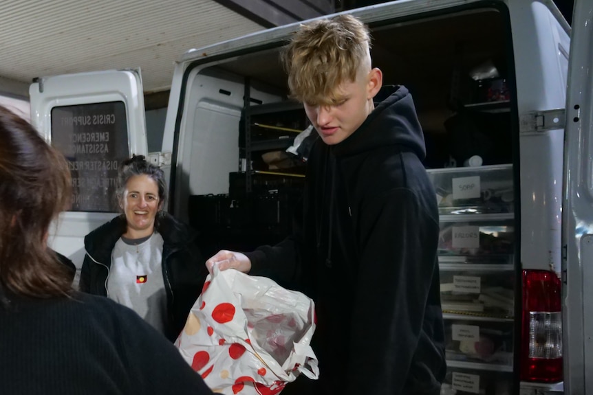 A tall teenage boy with a black hoodie fills a supermarket bag with donated goods. 
