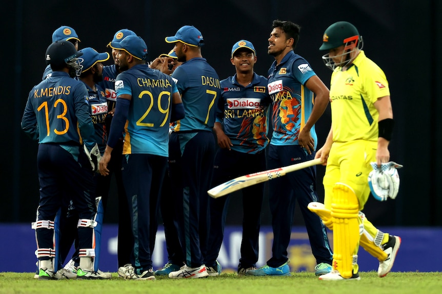 A cricket player wearing yellow holds his bat and walks off as other players in blue celebrate