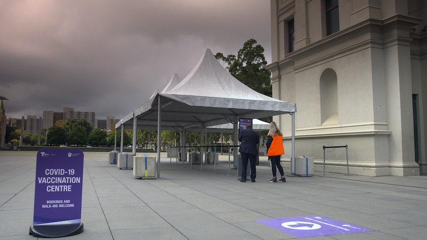 Two people stand ahead outside a vaccination centre. There are bollards and barriers for a queue but there is no one queuing. 