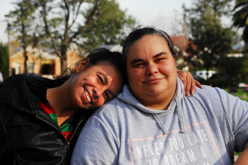 Tanya Day, smiling wearing a black jumper, rests her head on her daughter Belinda, smiling, wearing a grey jumper, in a park.